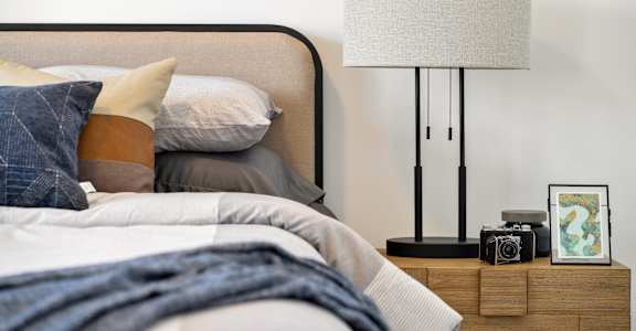 A bed with a blue blanket and a white pillow is in front of a lamp and picture frame.