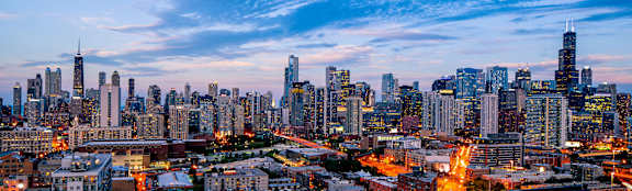A city skyline at dusk with buildings illuminated against a twilight sky.