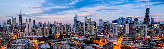 A city skyline at dusk with buildings illuminated against a twilight sky.