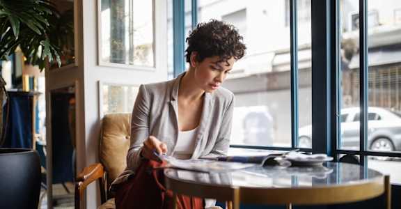 A woman engrossed in reading a magazine at a cafe table by a window, with a casual blazer over a white top, exemplifying an urban morning scene.