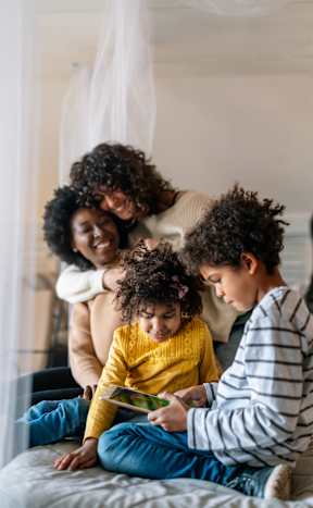 A woman and two children are sitting on a bed, with the woman leaning over the children as they look at a book together.