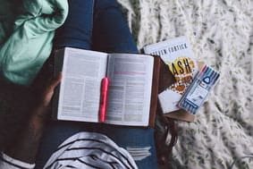 Open book with red marker resting on legs of person in jeans