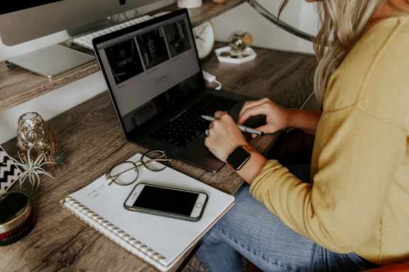 A person is sitting at a desk with a laptop, a smartphone, glasses, and a notebook.