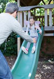 A child sliding down a green slide with a man pushing her.