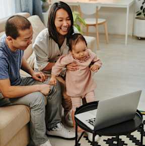 A family of three is sitting on a couch with a laptop on a small table in front of them.