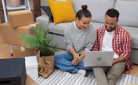 A man and woman are sitting on the floor with a laptop, surrounded by moving boxes and a plant.