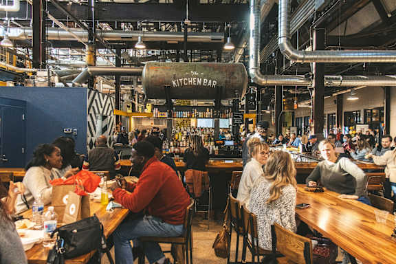 A group of people are sitting at tables in a restaurant with a sign that says Kitchen Bar.