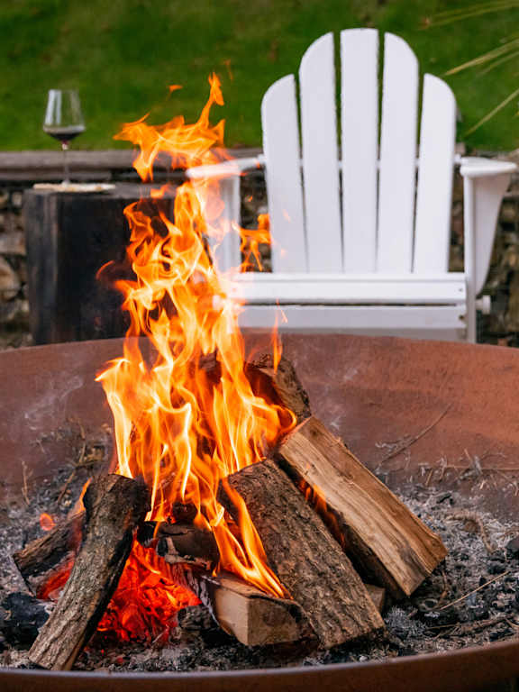 A white Adirondack chair is in front of a fire.