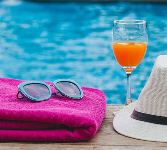 A glass of orange juice, a hat, sunglasses, and a towel are all placed on a wooden table by a pool.