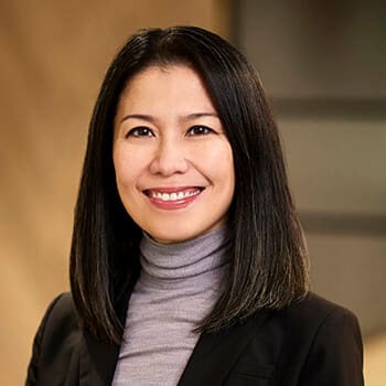 a woman with black hair and a grey turtleneck sweater smiles at the camera