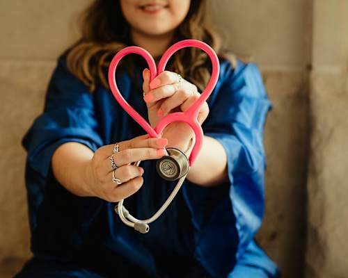 A person in a blue shirt is holding a pink heart-shaped object in one hand and a stethoscope in the other