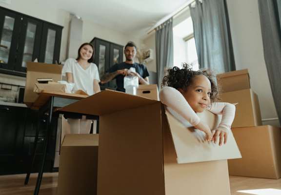 A family is moving into a new home, with cardboard boxes scattered around the room.