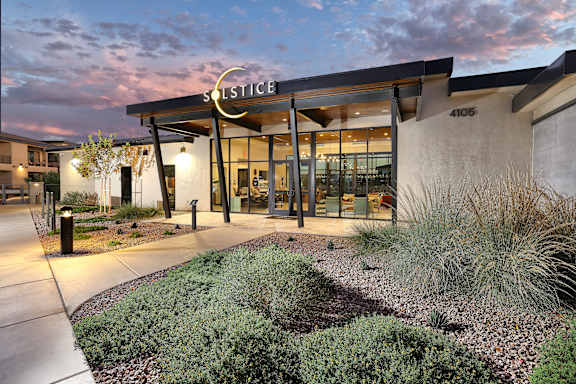 The image shows the front of a building named Solstice with a modern design, featuring a large glass entrance and a landscaped area in front.