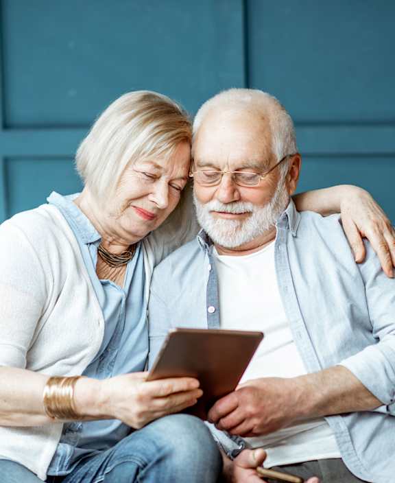 An elderly couple is sitting on a couch looking at a tablet.
