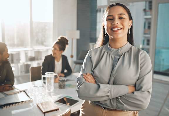 A woman stands with her arms crossed in front of a table with two other people sitting behind her.