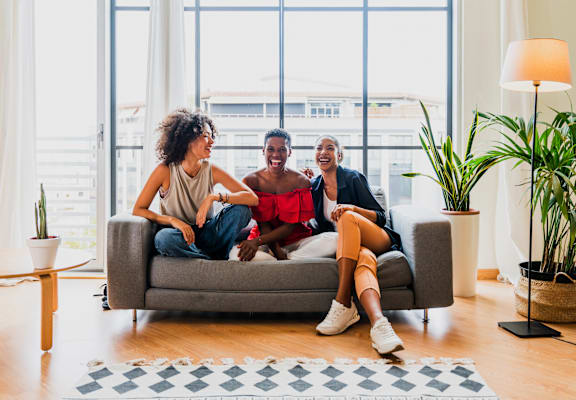 Three people are sitting on a couch in a bright room with large windows.