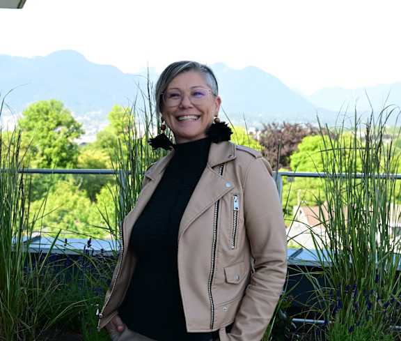 A woman in a tan jacket and black top stands in front of a car with a mountainous landscape in the background.