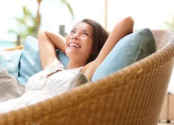 A woman is relaxing on a wicker chair with her arms behind her head.