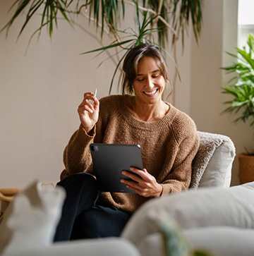 A woman in a brown sweater is sitting on a grey couch holding a tablet and a pen.