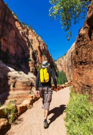 a man with a yellow backpack walks on a dirt path through a canyon