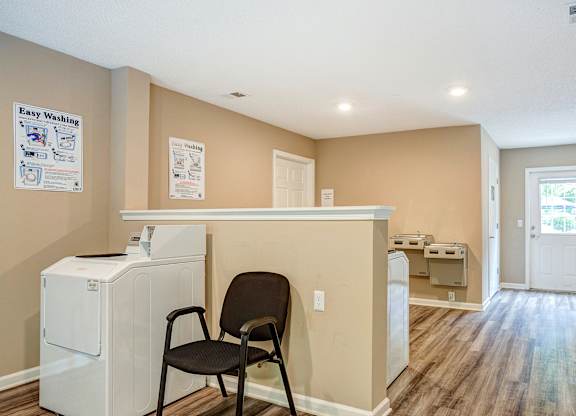 Community laundry room with beige walls, wood-style flooring, and bright lighting. The space features top-loading washing machines, stacked dryers, and a seating area with a black chair. Instructional washing signs are displayed on the walls, and a door with a window allows natural light to enter