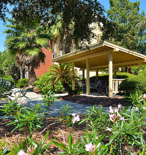 A small building with a porch is surrounded by greenery and flowers.