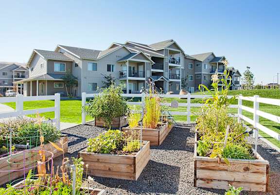 A white fence surrounds a garden with raised beds in front of a large house.