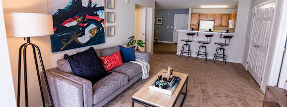 A grey couch with red and blue pillows sits in a living room with a painting on the wall.