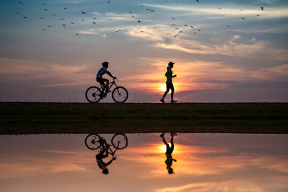 A person is riding a bicycle and another person is running on the grass with the sunset in the background.