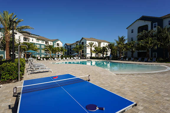 A blue table tennis court is surrounded by palm trees and buildings.