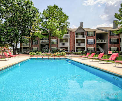 A large swimming pool in front of a building with red lounge chairs on the side.