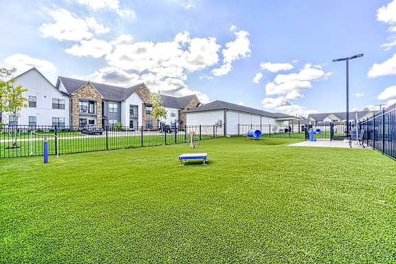 A grassy field with a bench and a fence in the foreground and houses in the background.