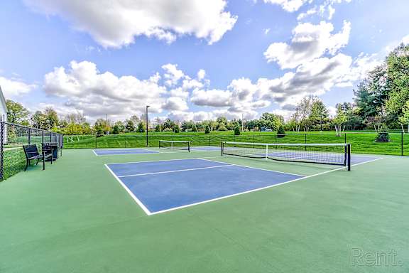A tennis court with a blue top and white lines, surrounded by a green fence and trees.