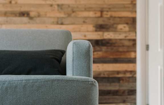 A grey couch with wooden legs sits in front of a wooden wall.