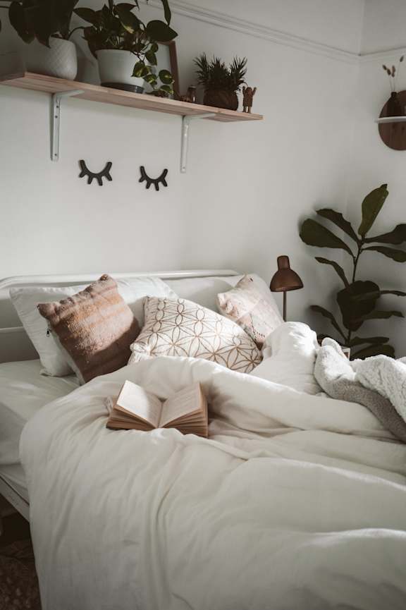 A cozy bedroom with a bed, pillows, a book, and plants.