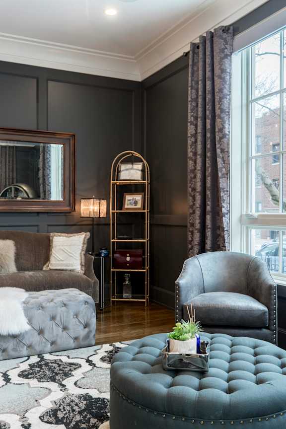 A grey tufted ottoman sits in front of a grey chair in a room with dark walls.