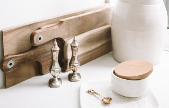 Cutting board on white counter. White vase with wooden spoons. 