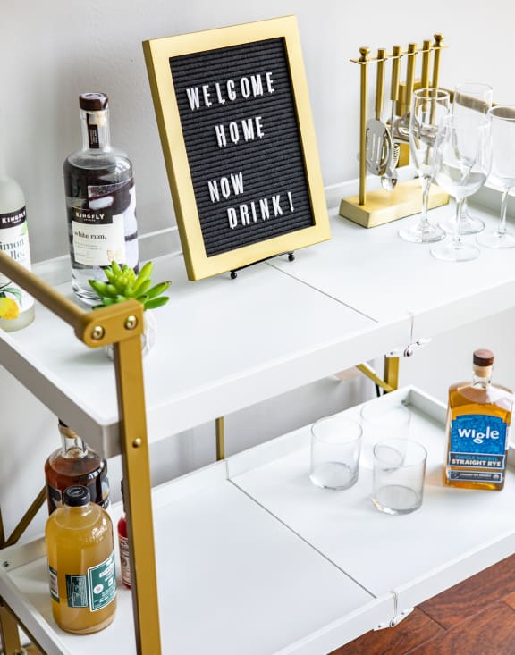 a white and gold bar cart with glasses on it and a sign that says welcome home