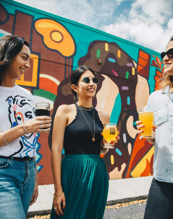 three women standing in front of a mural drinking beer