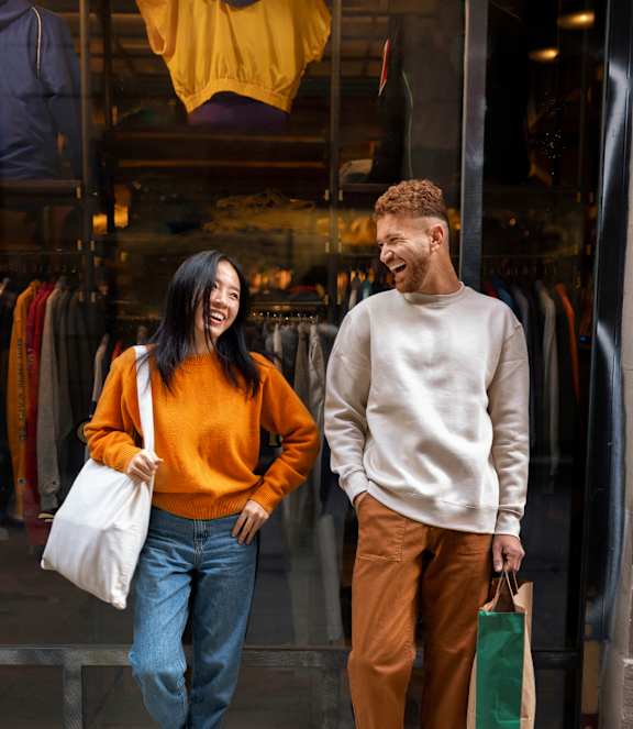 A man and a woman are standing outside a clothing store, both holding shopping bags.