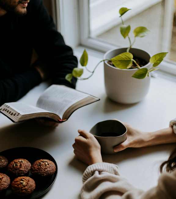 A person is reading a book while holding a cup and there is a plate of food on the table.