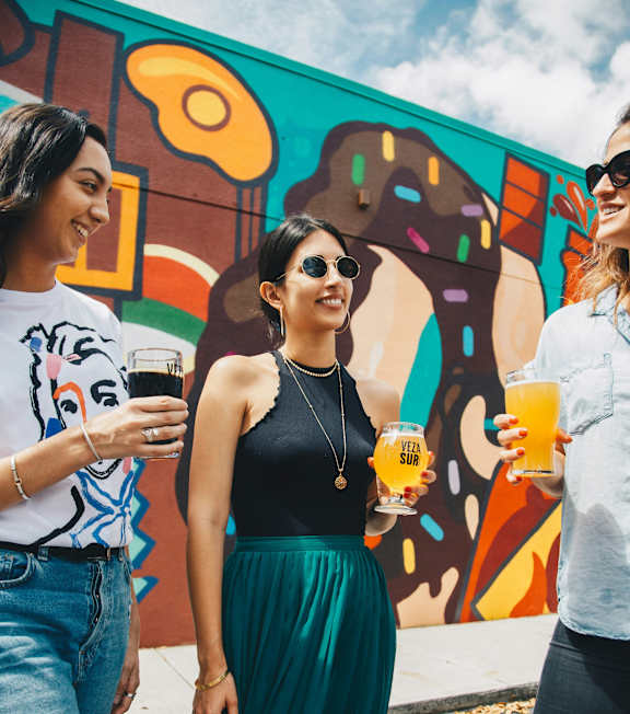 Three friends are standing in front of a colorful mural, each holding a drink.