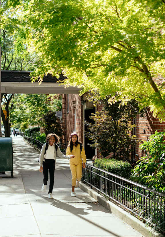 Two women walking down a sidewalk under a tree.