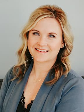 A woman with blonde hair wearing a grey blazer and a black lace top.