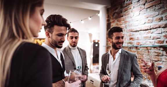 A group of well-dressed individuals at a social gathering, with a focus on three men engaging in conversation while holding drinks, against a backdrop of a cozy interior with exposed brick walls.