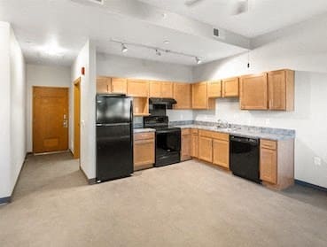 an open kitchen and living area with ample wood cabinetry, black appliances, and overhead lighting at Muskego School Apartments