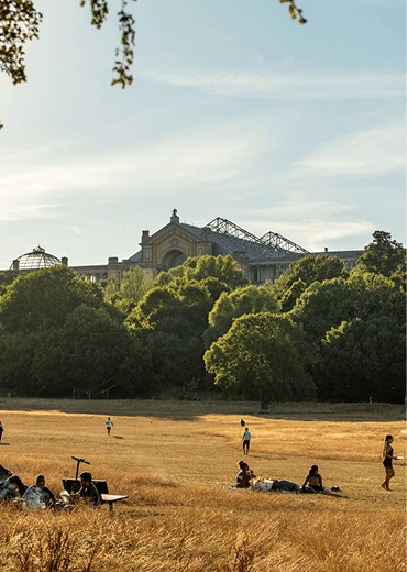 A group of people are sitting and standing on a grassy field with a large building in the background.