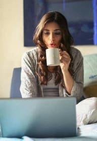 a woman sitting on a bed with a cup of coffee looking at a laptop