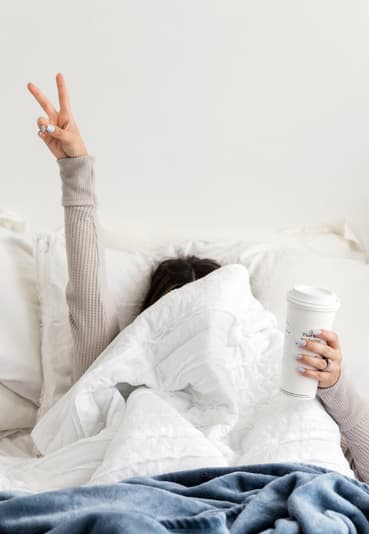 Person in bed holding coffee cup with fingers in peace sign