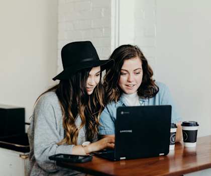 Two women are sitting at a table looking at a laptop.